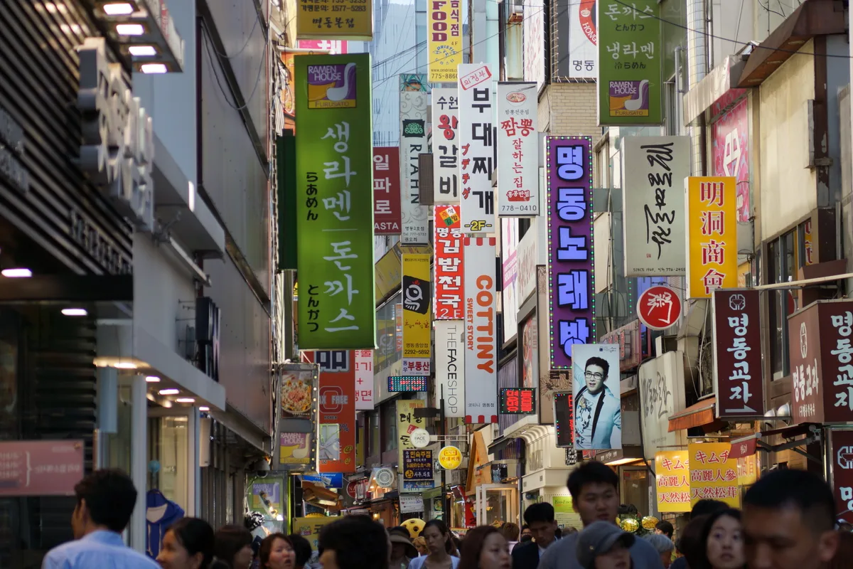 A crowded Myeongdong shopping street in Seoul lined with stacked restaurant and shop signs.