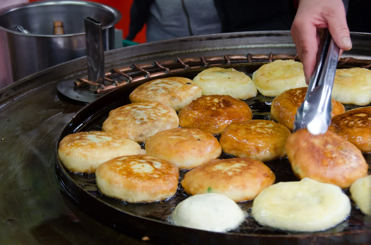 A street vendor uses tongs to turn hotteok, sweet filled pancakes, cooking on a round griddle.