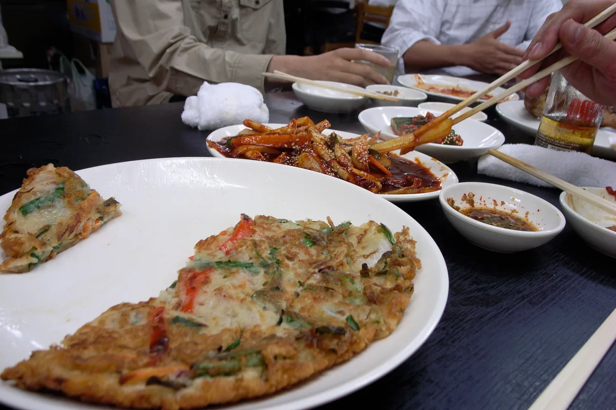A savory Korean pancake (pajeon) with scallions and vegetables, sliced on a plate.