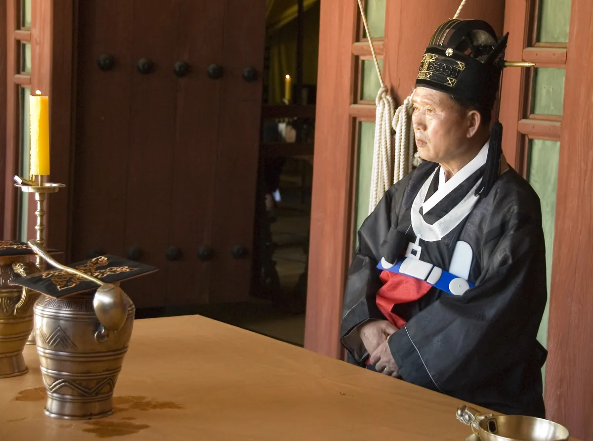 An official in formal Jongmyo Jerye ceremonial dress standing before a ritual altar with offering vessels and a lit candle.