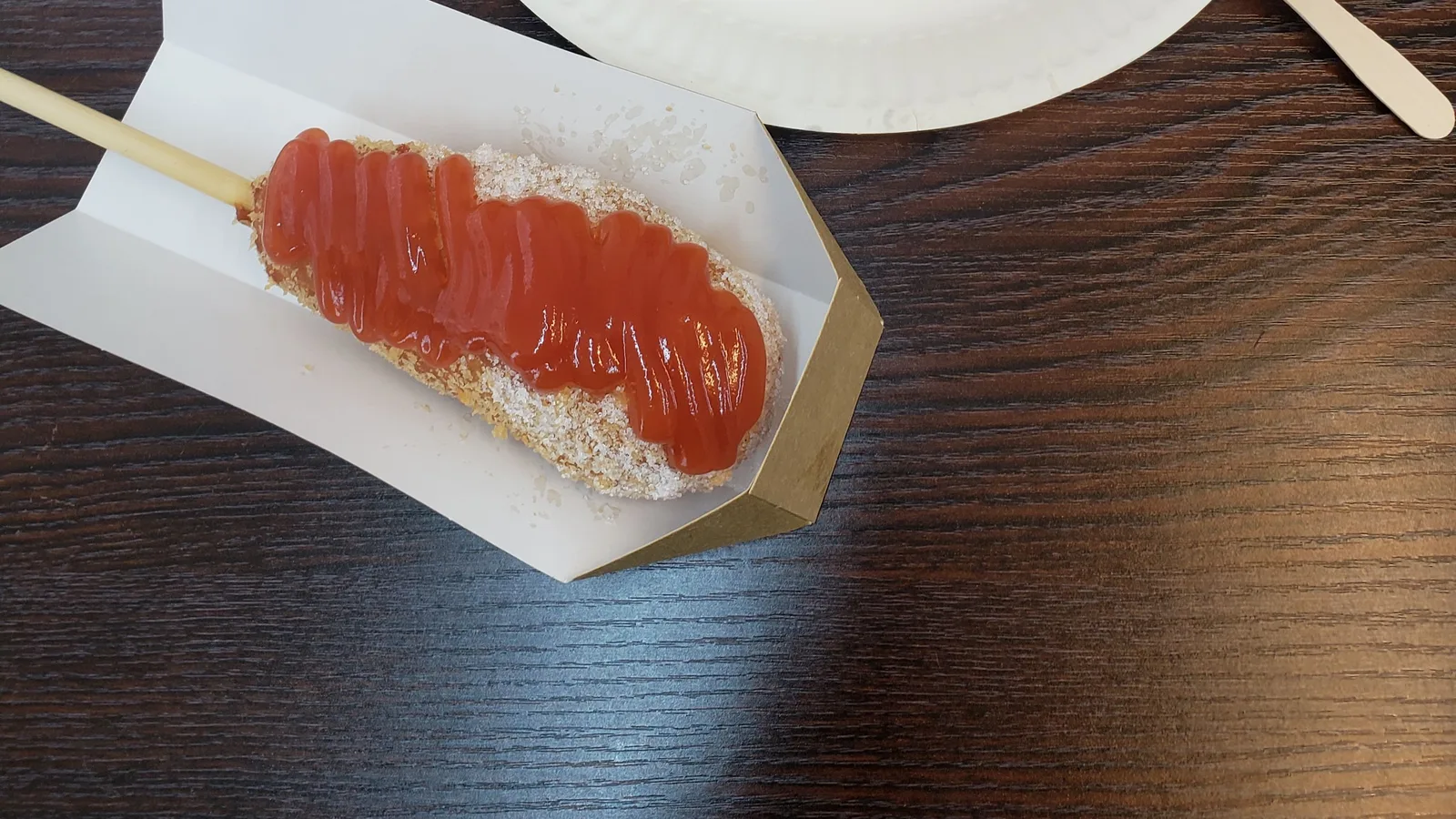 A Korean corn dog coated in a crunchy breaded exterior and drizzled with ketchup, served on a wooden stick.