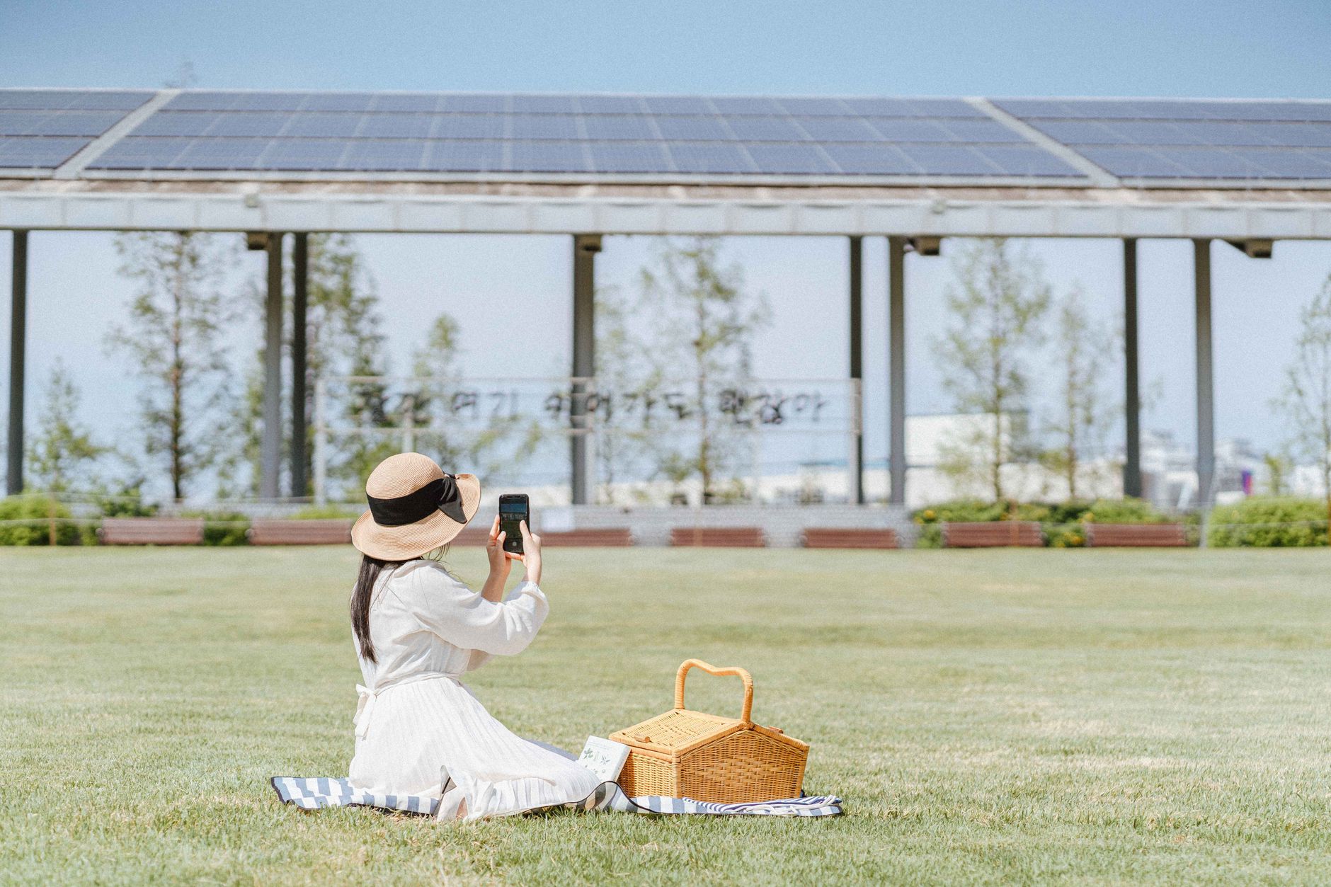 A woman in a white dress and sun hat takes photos during a picnic in a sunny park.