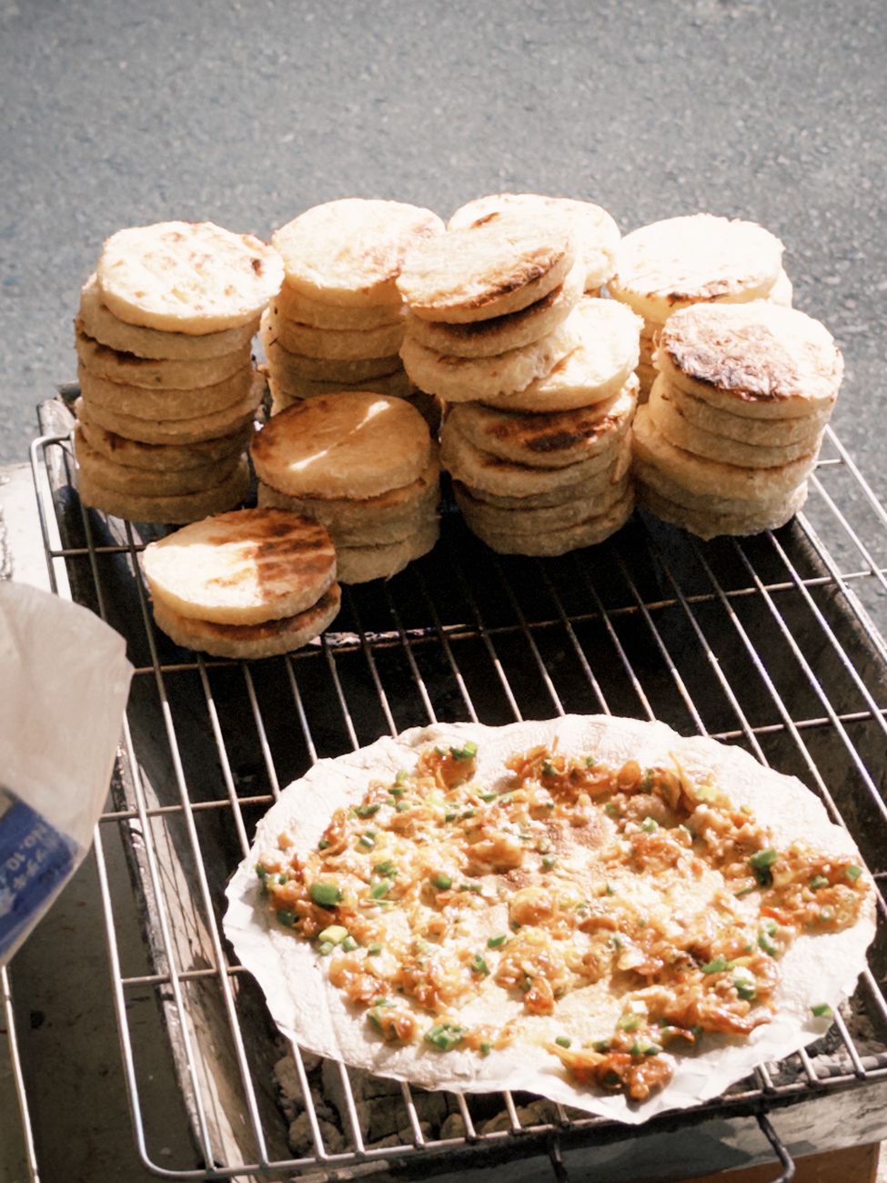 Stacks of flatbreads on a grill with one topped with vegetables in outdoor sunlight.
