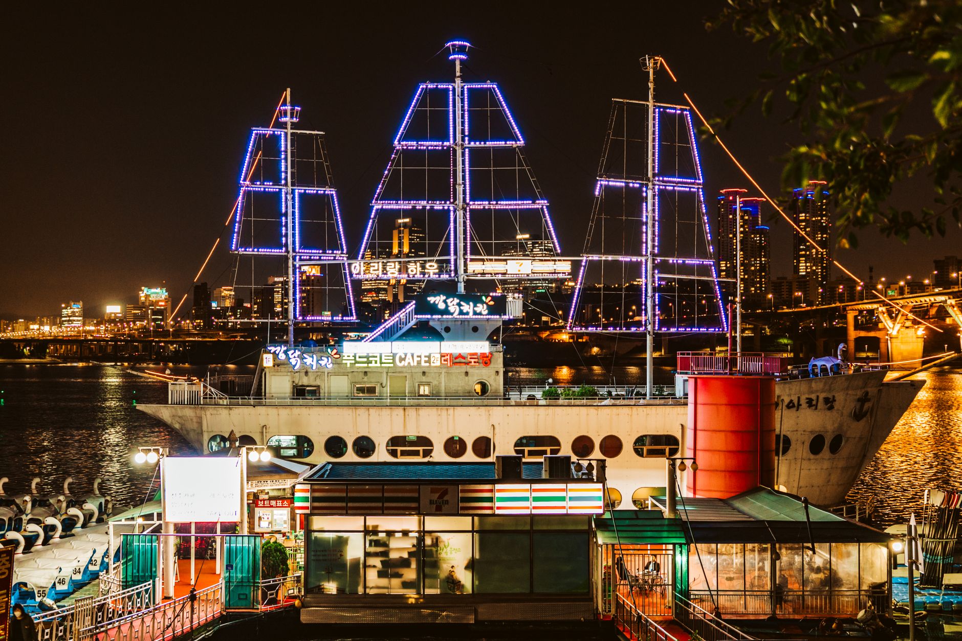 A brightly lit ship cafe moored on the Hangang River with city skyline in the background at night.