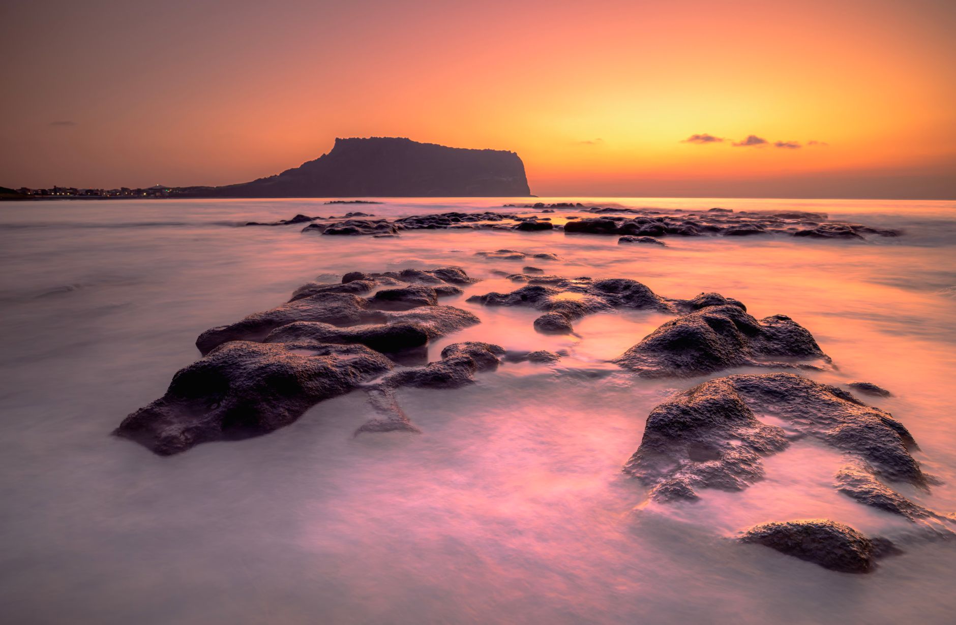 Stunning sunrise over Seongsan Ilchulbong Peak, Jeju Island, South Korea, featuring smooth ocean and rocky foreground.