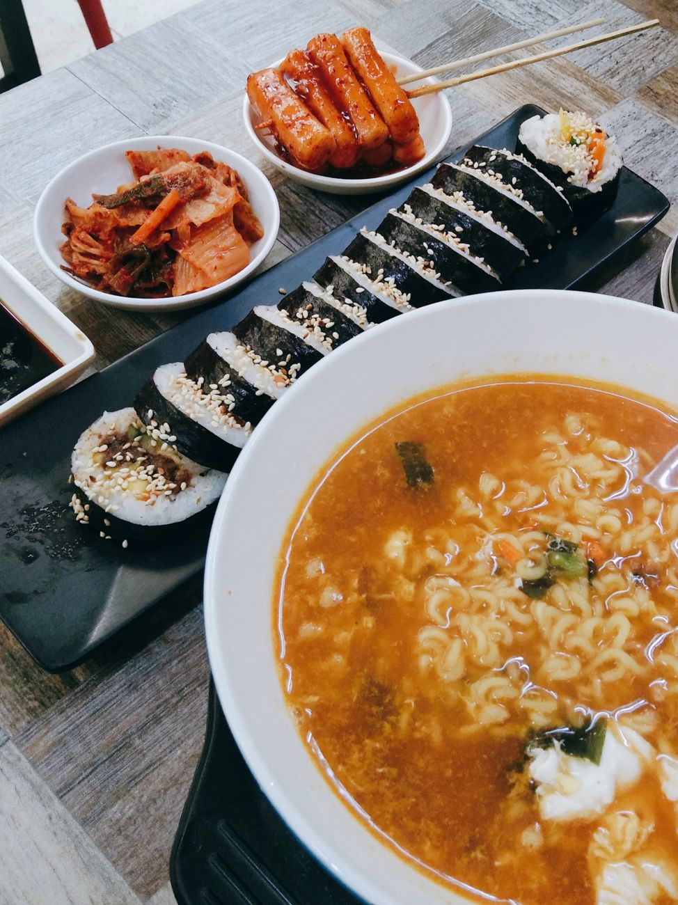 Delicious Korean meal featuring hot ramen, gimbap, kimchi, and tteok-kkochi on a wooden table.