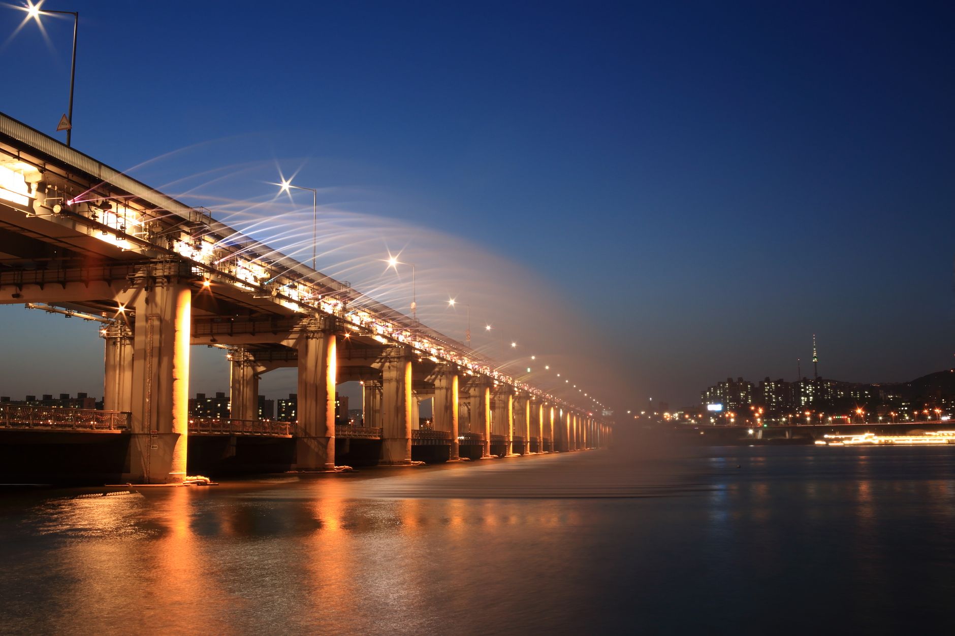 Beautiful night view of a brightly lit bridge over a calm river with city lights in the background.