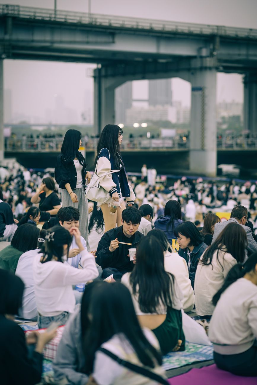 A vibrant crowd gathered outdoors under a bridge in Seoul, enjoying a lively event during the day.