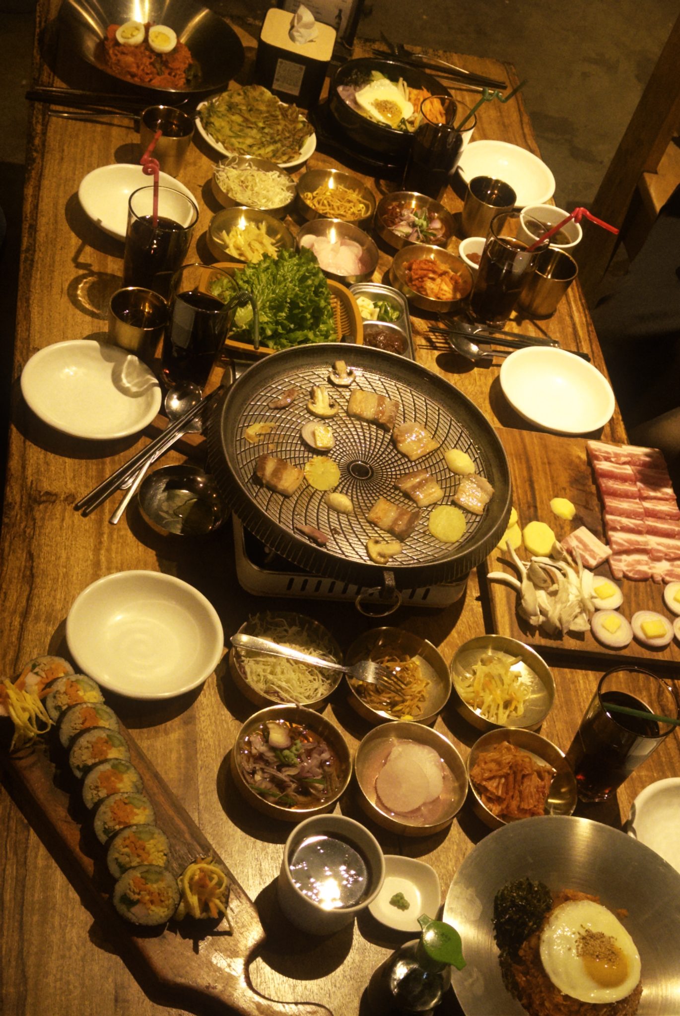 An overhead view of a full Korean BBQ spread on a wooden table featuring a round charcoal grill with meat and mushrooms cooking, raw pork belly slices…