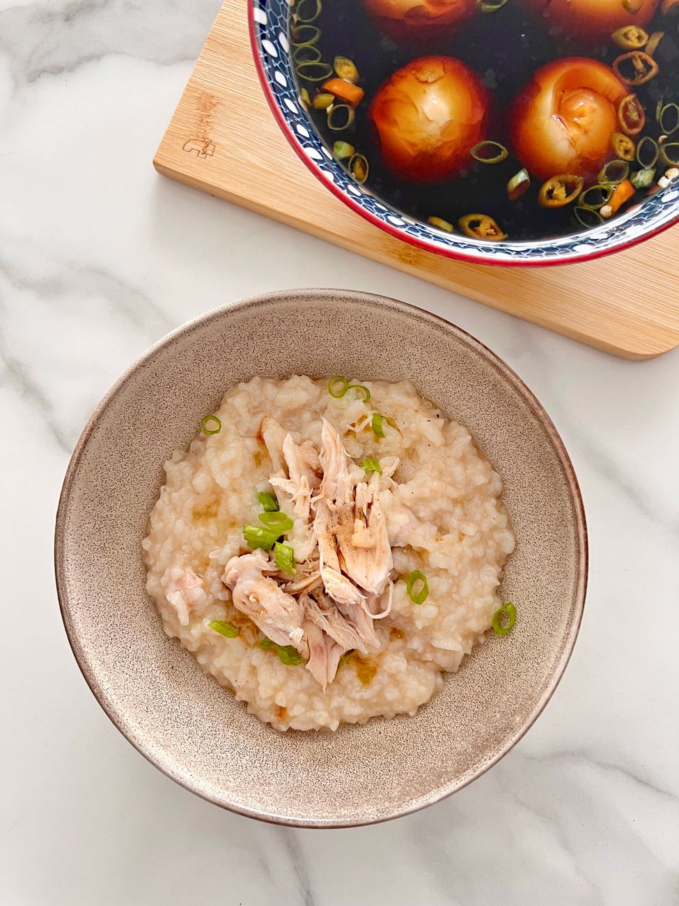 A bowl of creamy rice porridge topped with shredded chicken and green onions, alongside a dish of soy-marinated eggs.