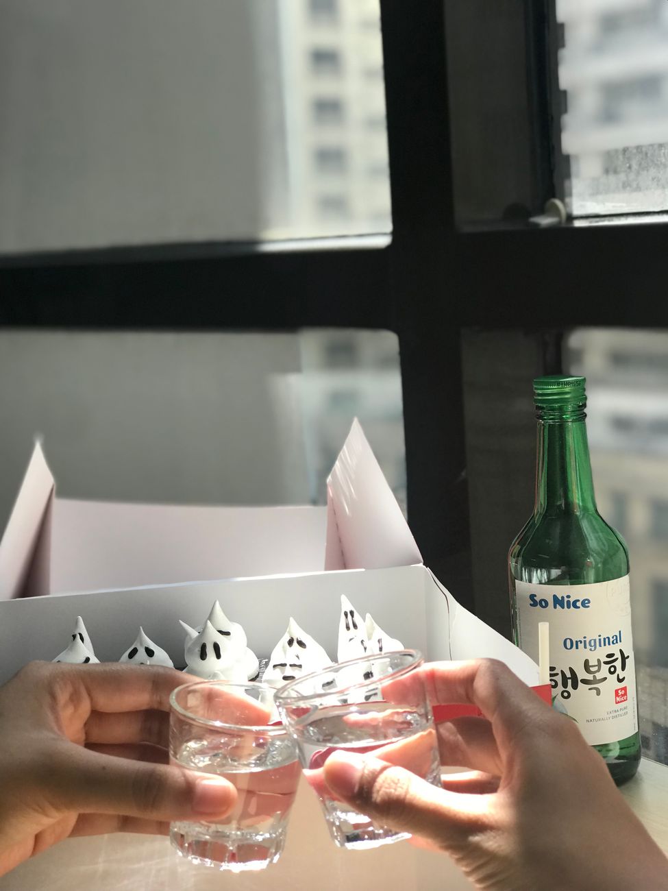 Hands clinking glasses of soju with ghost cupcakes by window.