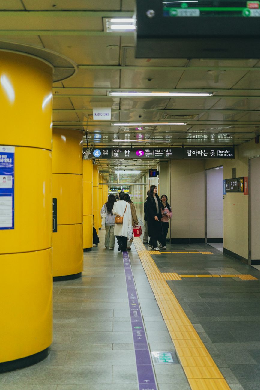 Seoul metro station scene depicting commuters walking amidst vibrant yellow pillars.