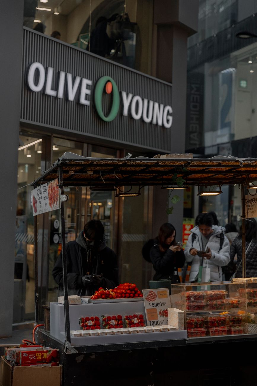 Street vendors selling produce outside Olive Young store in city setting.
