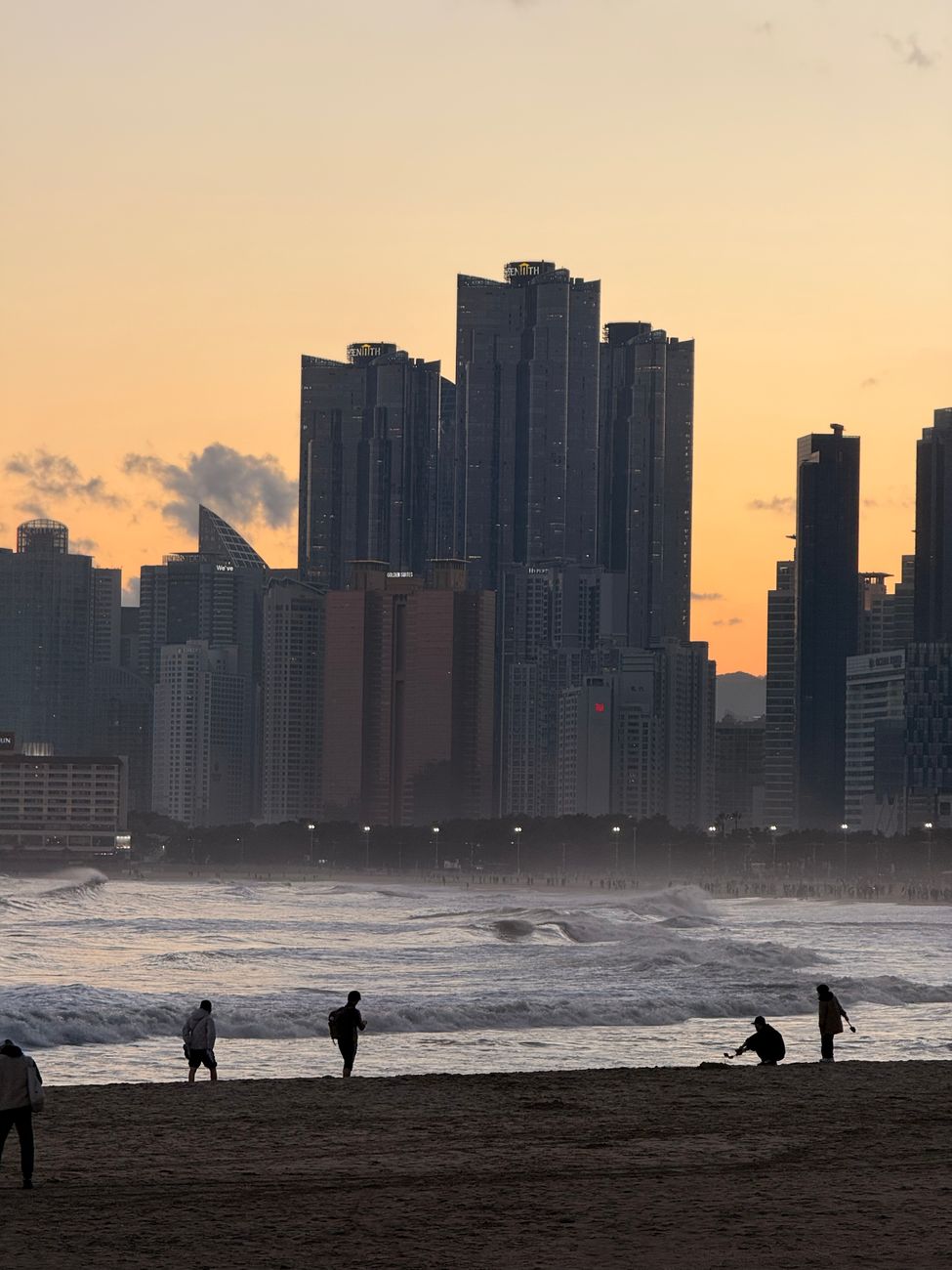 Silhouetted figures on Haeundae Beach against the backdrop of Busan's skyline at sunset.