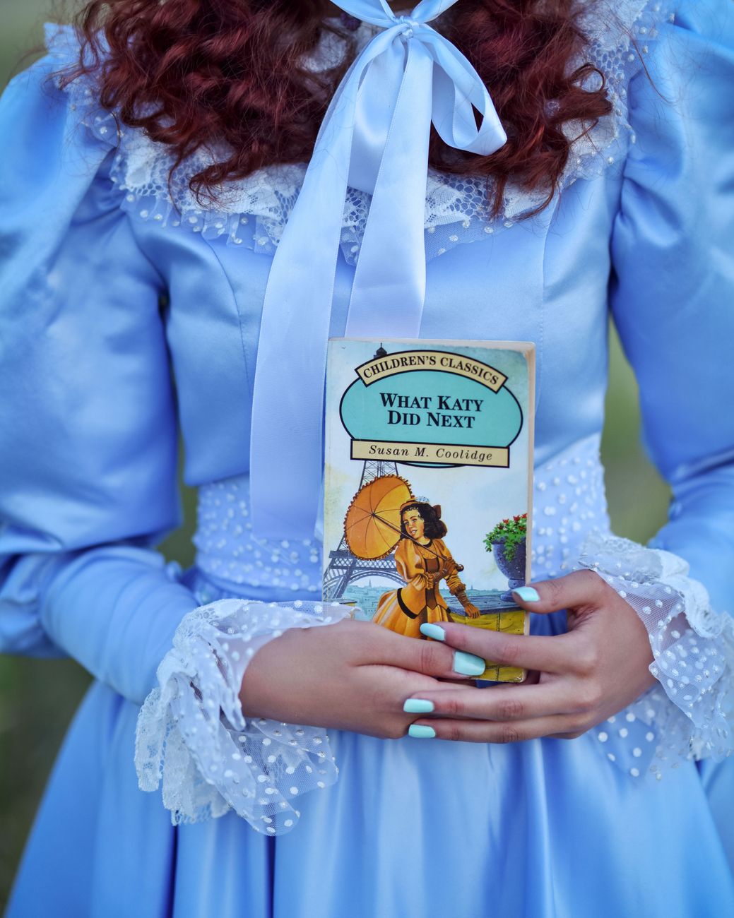 Woman in vintage blue dress holding 'What Katy Did Next' book illustrating classic style.