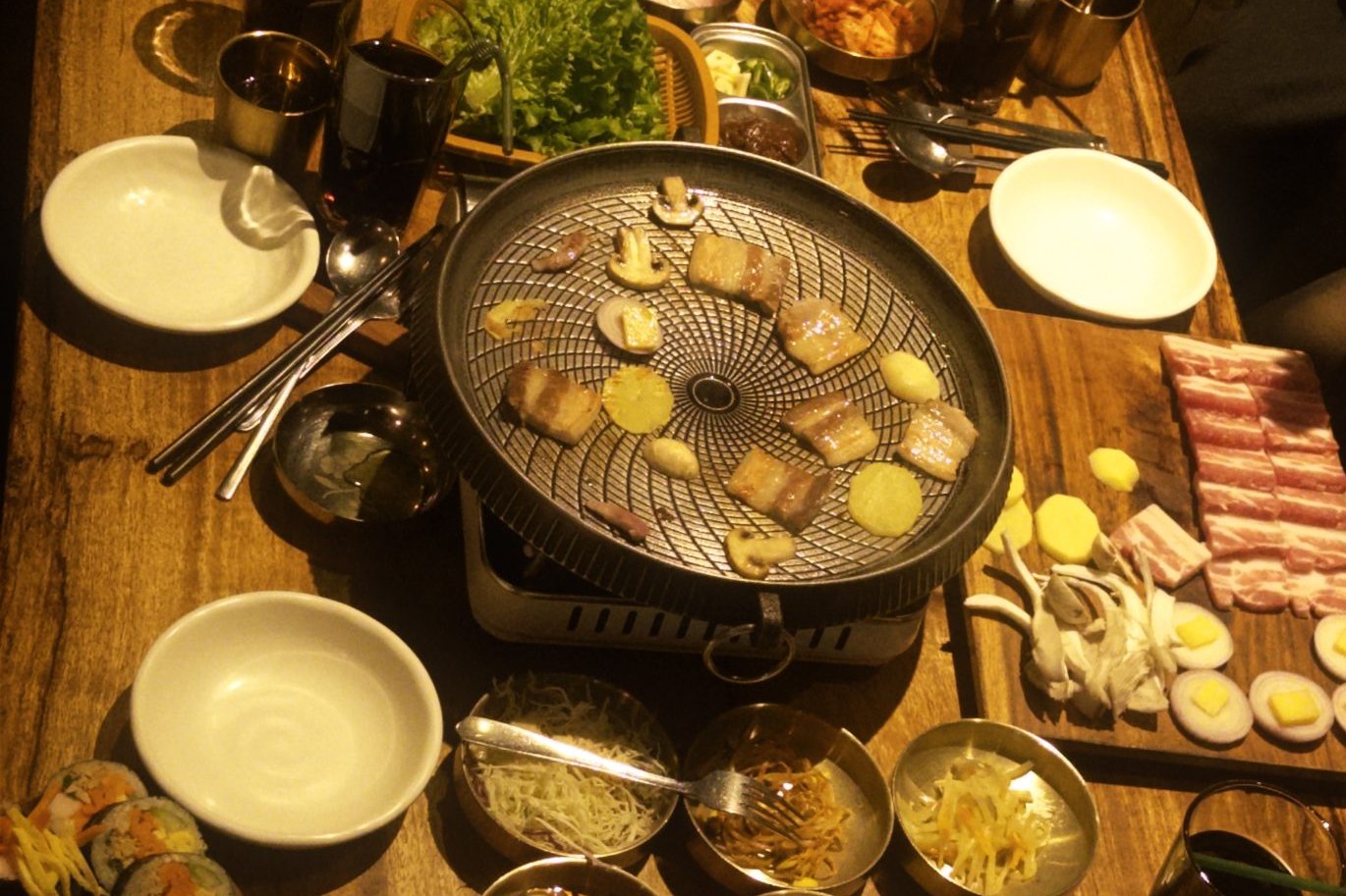 An overhead spread of a Korean BBQ meal: a round grill in the center with pork belly cooking, surrounded by lettuce wraps, gimbap, and many banchan side dishes.