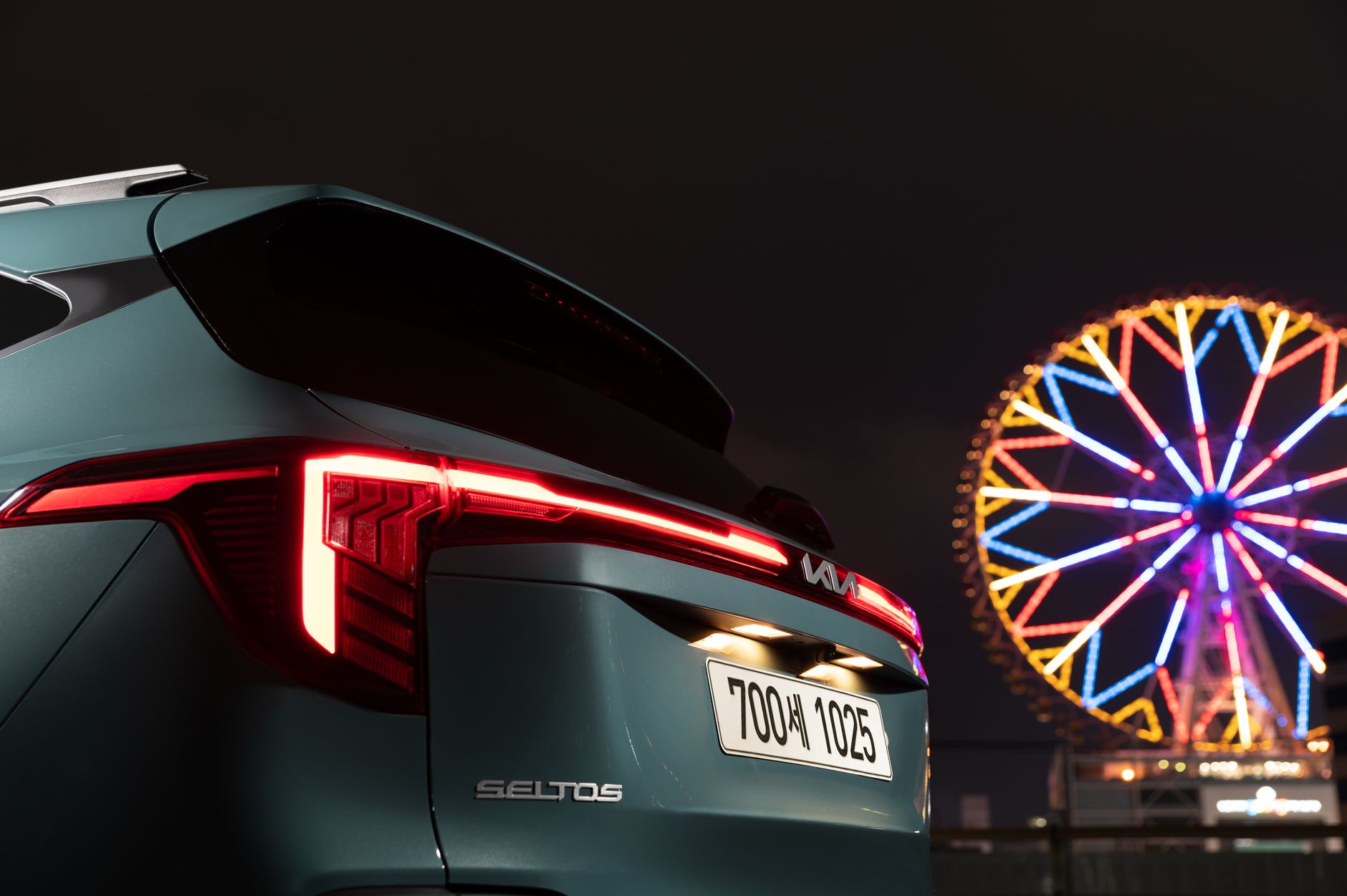 A Kia Seltos SUV parked at night with a colorful ferris wheel in the background.