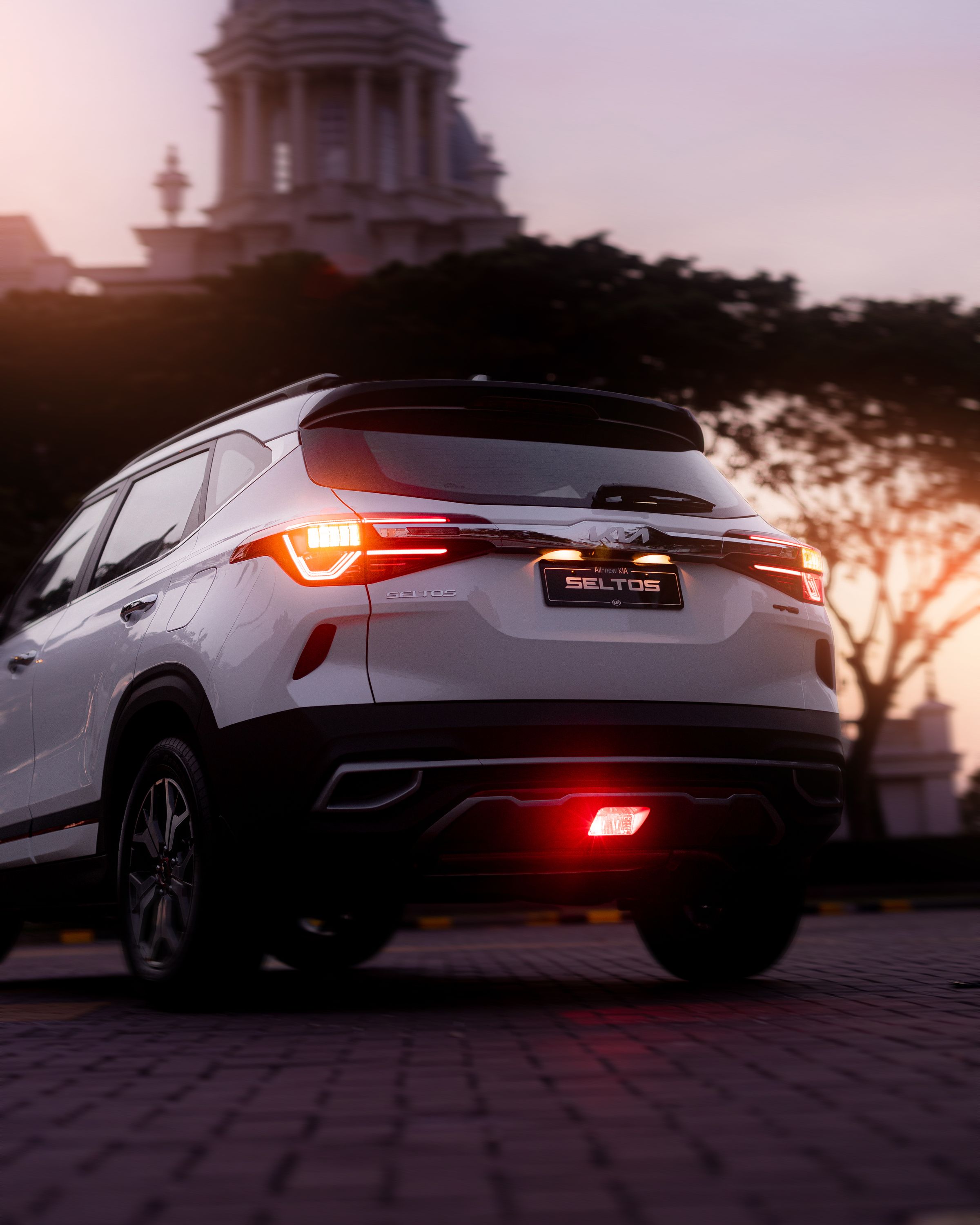 A white Kia Seltos SUV seen from the rear, taillights illuminated, against a city skyline at sunset