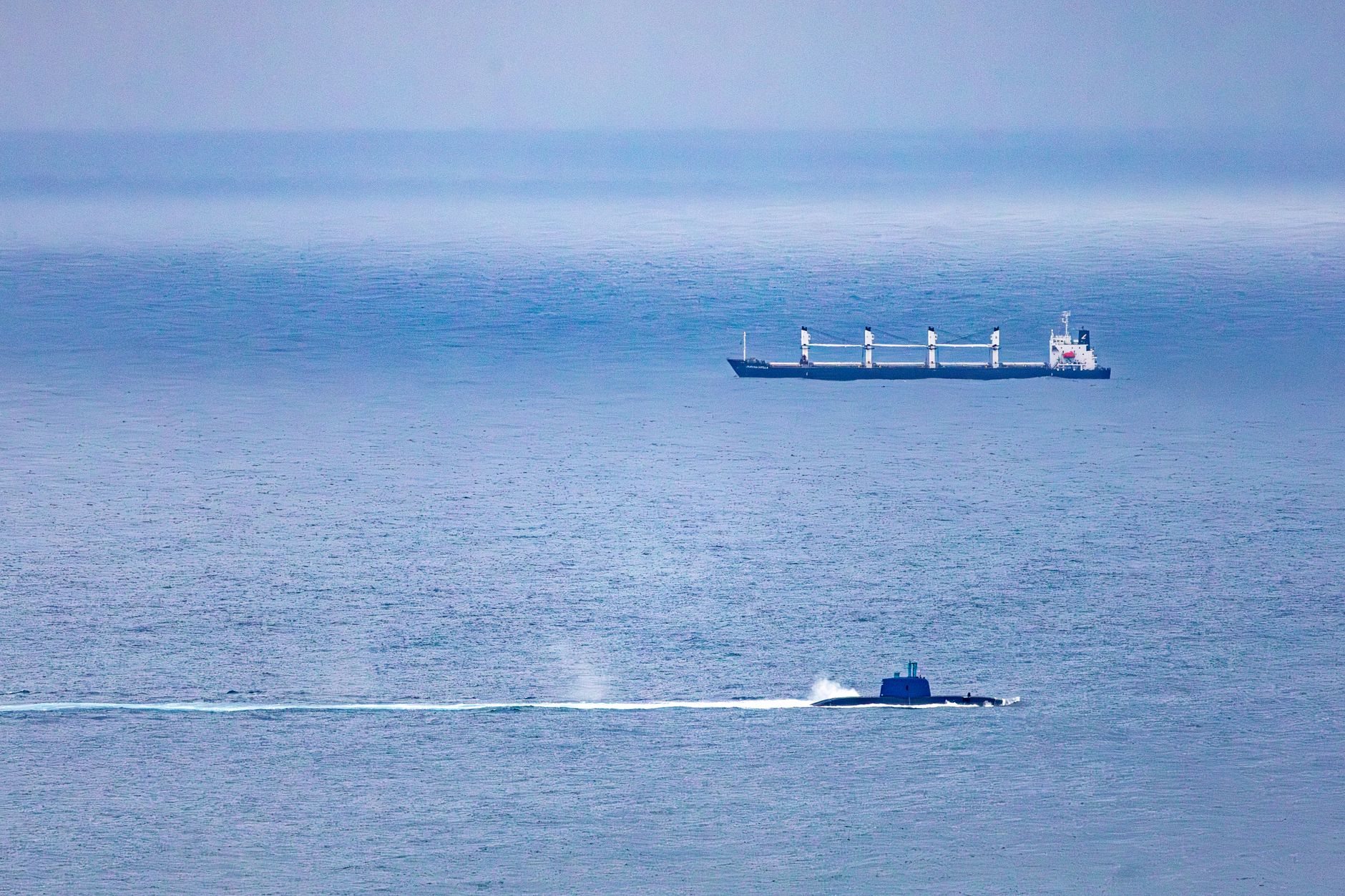 A cargo ship and submarine navigate the vast, open ocean under a clear sky.